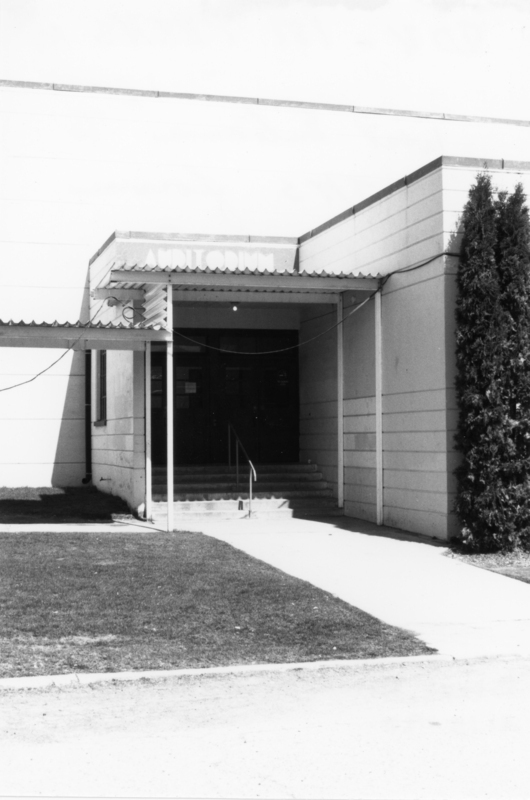 A building entrance with a canopy labeled "Auditorium," featuring steps leading to double doors. There is a pathway and a small area of grass in front, with a tall shrub on the right side.