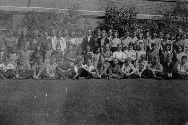 A large group of children and a few adults posed together on a grassy area in front of a building. Some are sitting cross-legged in the front row, while others stand behind them. The background includes a wall with windows and some trees or bushes.