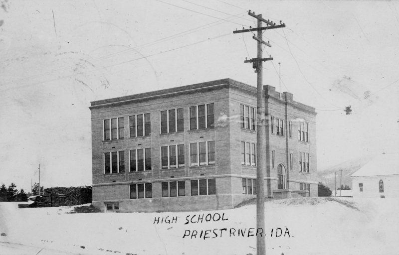 A large, rectangular brick building with many windows, labeled as a high school. It is surrounded by snow, and there is a utility pole with wires in the foreground. Another smaller building is visible to the right. The text reads: "HIGH SCHOOL PRIEST RIVER, IDA."