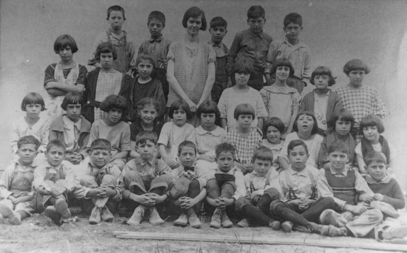 A group of children and one adult standing and sitting together in rows, likely forming a class or group photo. The adult stands in the middle of the top row, with children of various ages positioned around them. The children have a range of expressions and are dressed in simple clothing.