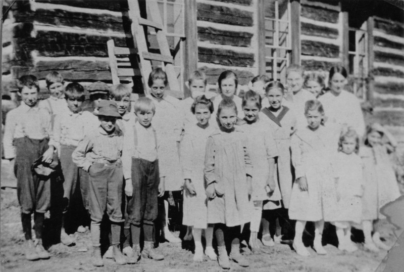 A group of children standing in front of a wooden building with windows and ladders. They are wearing a variety of clothing, including dresses and pants with suspenders. Some children are smiling, and a few have their hands in their pockets.