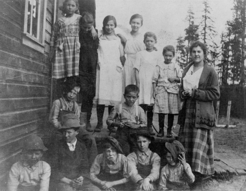 A group of children and one adult standing and sitting on a porch. The children are dressed in various clothing, including dresses and overalls. Trees are visible in the background.