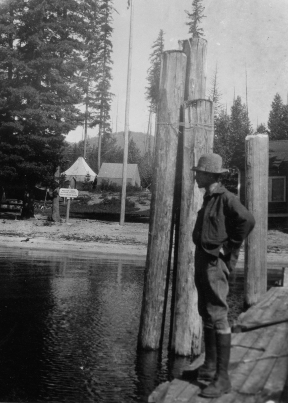 A person stands on a wooden dock overlooking a body of water. Nearby, there are tall wooden posts. In the background, a campsite is visible with tents and tall trees surrounding the area. Sign in background reads: "Beaver Creek Ranger Station Dist. Ranger HDQS".