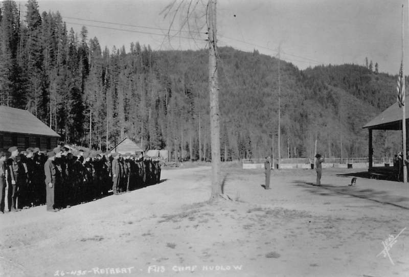 A group of people standing in rows near a building in a forested area. An individual stands at attention in front of them, possibly leading an event. There are structures in the background and trees in the distance. Text near the bottom reads: "26-435-RETREAT F113 CAMP HUDLOW".