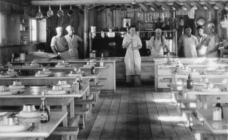 A group of people, mostly wearing aprons and hats, stand in a kitchen with a large stove and hanging pots in the background. Tables with set plates and bottles are arranged in the foreground.