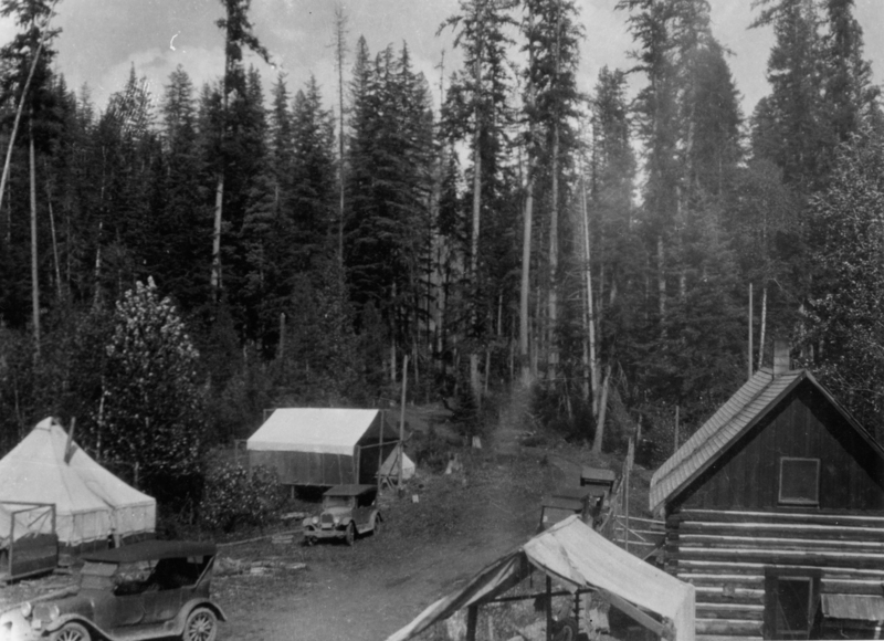 A cabin and several tents situated among tall trees, with two vintage cars parked on a dirt path.