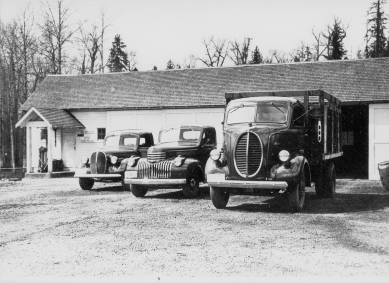 Three vintage trucks are parked in front of a long building with a sloped roof. A person is standing under the porch at the entrance of the building. Trees are visible in the background. The sign on the building reads "NO SMOKING. STOP YOUR MOTOR."