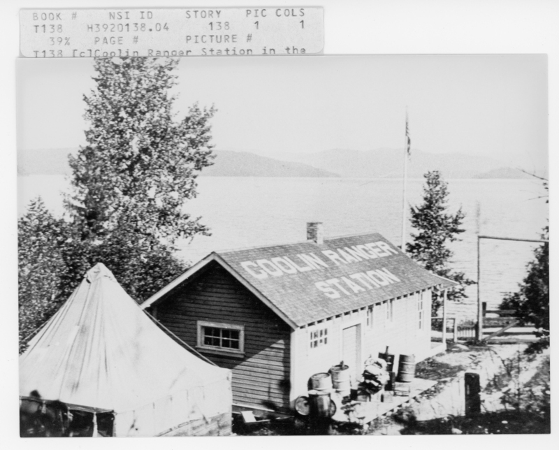 A wooden building labeled "Coolin Ranger Station" is situated near a body of water with trees surrounding it. There is a tent nearby and a flagpole standing next to the building. Text reads: "- Text at the top reads: - "BOOK # NSI ID STORY PIC COLS" - "T138 H3920138.04 138 1" - "39% PAGE # PICTURE #" - "1 138 (E)(Coolin_ Ranger Station in the" - Text on the building's roof reads: - "COOLIN RANGER STATION".