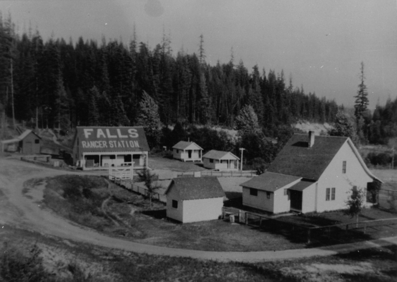A group of small buildings situated in a rural area surrounded by tall trees. One building has a large sign on the roof that reads "FALLS RANGER STATION." The buildings are connected by dirt paths, and there is a small fenced area in front of one structure.