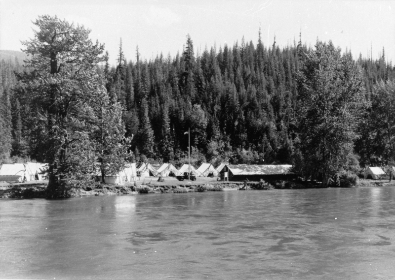 A row of tents and a wooden building are situated along the bank of a river. Large trees are positioned in front of a dense forest in the background. A vehicle is parked among the tents.