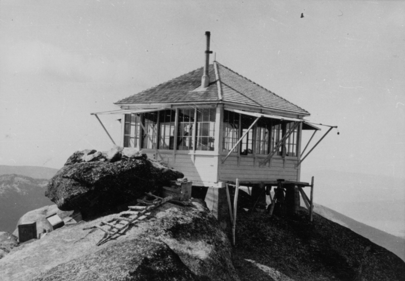 A small wooden structure with a chimney is perched on top of a rocky hill, supported by wooden beams. A ladder made of branches is leaning against the rocks near the building. There are mountains in the background.