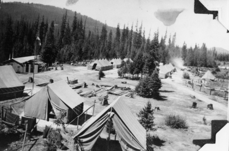 A group of tents arranged in a forested area with tall trees in the background. There are piles of logs and some structures scattered throughout the site. An American flag is visible on a pole near one of the tents.