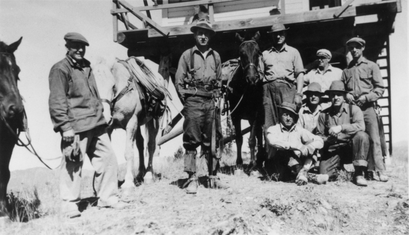 A group of men standing and sitting on a rocky hillside with two horses nearby. They are gathered near a building with wooden beams. Some men are wearing hats and outdoor clothing.