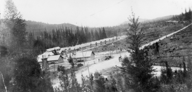 Rows of small wooden buildings in a clearing surrounded by forested hills. Dirt roads run between the structures, with a few trees and small clearings around the area. Some vehicles and people are visible near the buildings.