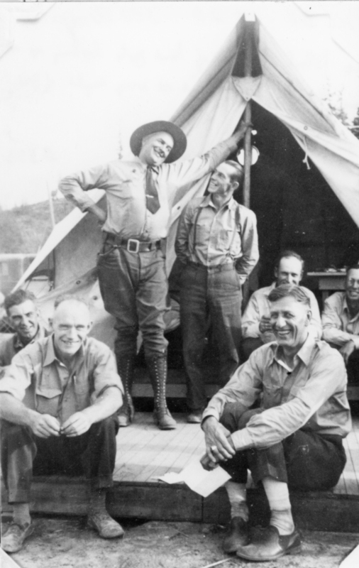 A group of eight men gathered in front of a canvas tent. Two are standing on the steps, one of whom is pointing upwards, while the rest are seated or crouching on the ground and steps, smiling and relaxed. They wear a mix of shirts, pants, and boots, suggesting a casual outdoor setting.