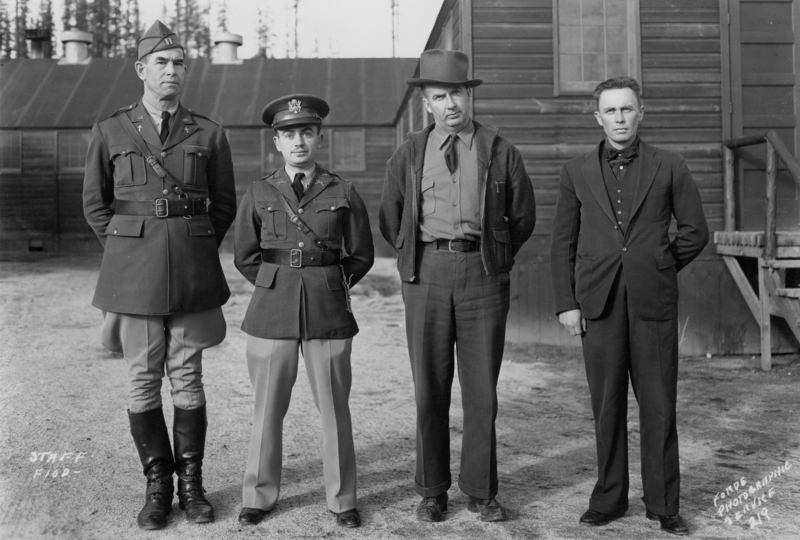 Four men standing side by side outdoors, with wooden buildings in the background. Two are in military uniforms with caps, one wears a cowboy hat and casual jacket, and the last is dressed in a suit with a bow tie. Near the bottom left, text reads: "STAFF F102". Near the bottom right, text reads: "Colony Photographic @ 214 719".