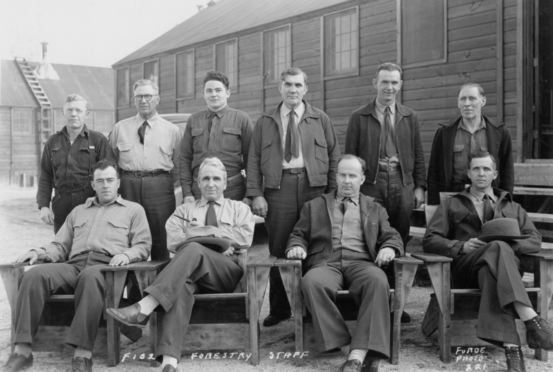 A group of ten men is posed in two rows in front of a wooden building. The front row consists of four men sitting in chairs, while six men stand behind them. The men are dressed in collared shirts and jackets. Text at the bottom reads: "F.102 FORESTRY STAFF." On the lower right, it reads: "FORDE PHOTO A 21."