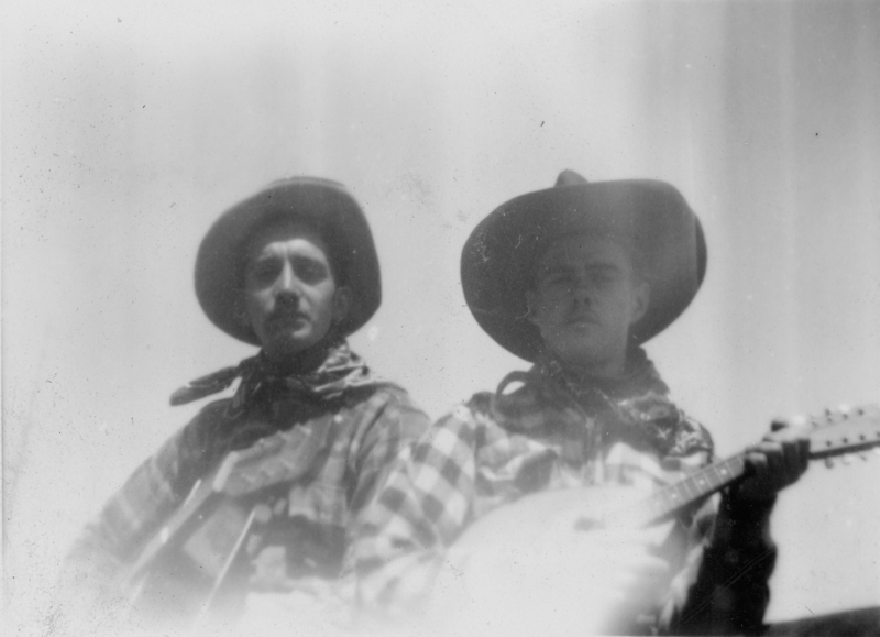 Two men wearing cowboy hats and plaid shirts, one holding a stringed instrument similar to a mandolin.