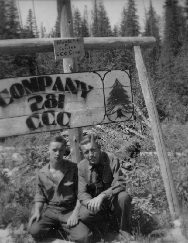 Two men sitting on the ground in front of a wooden sign with faded letters and a tree illustration. Dense forest is visible in the background. The sign near the top reads: "HUNTERS BE CAREFUL CCC Camp". The larger sign reads: "COMPANY 281 CCC".
