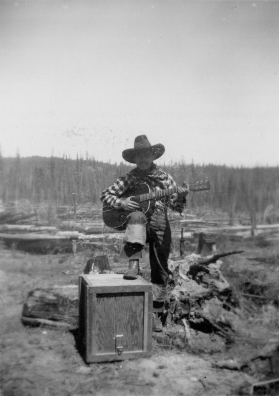 A person wearing a wide-brimmed hat and plaid shirt stands outdoors, playing a guitar with one foot on a wooden box. The background features a wooded landscape with scattered logs and trees.