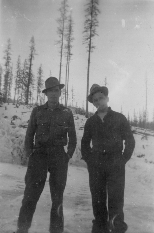 Two men standing in a snowy landscape with tall, thin trees in the background. They are wearing hats and jackets, with their hands in their pockets.