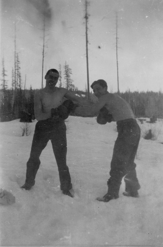 Two men are boxing in the snow. They are shirtless, wearing trousers and boxing gloves. Tall, bare trees stand in the background, and snow covers the ground around them.