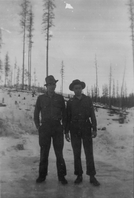 Two men wearing hats and dark clothing stand on a snowy landscape with tall, sparse trees in the background.