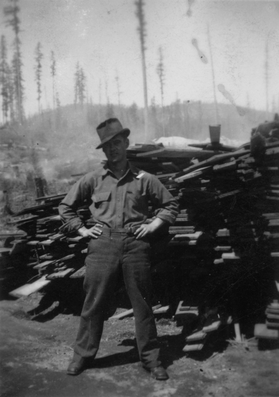 A man wearing a hat and work clothes stands with his hands on his hips in front of a pile of wooden planks, with trees in the background.