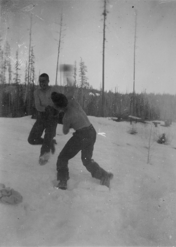 Two people are playfully sparring or boxing in a snowy landscape. They are both wearing boots and pants, with bare torsos. In the background, there is a forest with tall, thin trees.