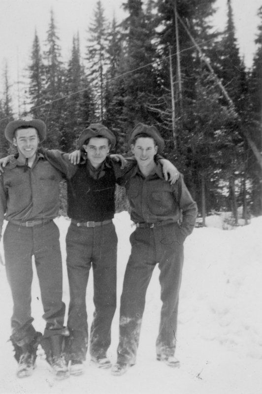 Three men standing together in a snowy landscape, each wearing hats and outdoor clothing. They have their arms around each other in a friendly pose, with tall trees in the background.