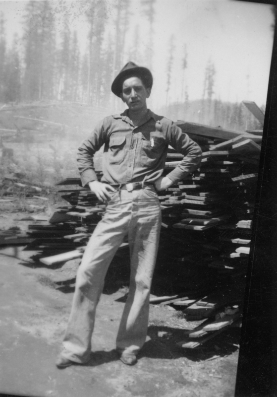 A man stands in front of a stack of lumber, wearing a buttoned shirt, loose pants, and a hat. He has his hands on his hips and there are trees in the background.