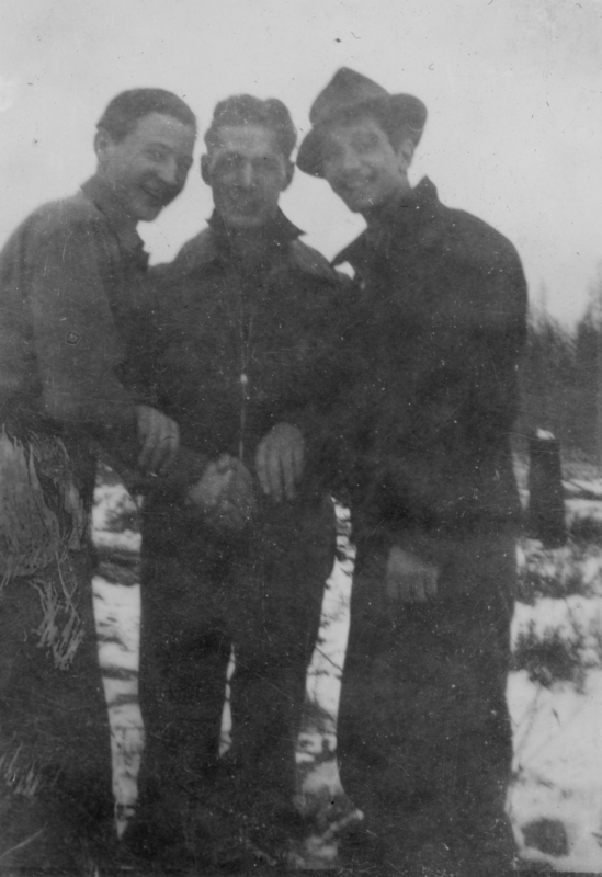 Three men standing closely together outdoors, with two of them wearing hats, against a backdrop of snow and trees.