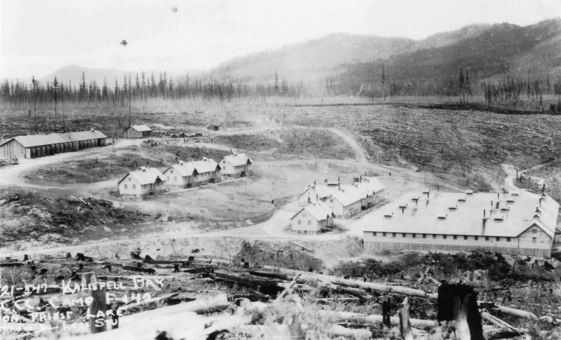 A series of wooden buildings are set in a clearing surrounded by hills and sparse forest. A network of dirt roads connects the structures. The landscape appears rugged and partially deforested. Words on the lower left hand corner of the screen read "21-547- Kalispell Bay... CCC Camp F-142...On Priest Lake... Photo by Leo's Studio".