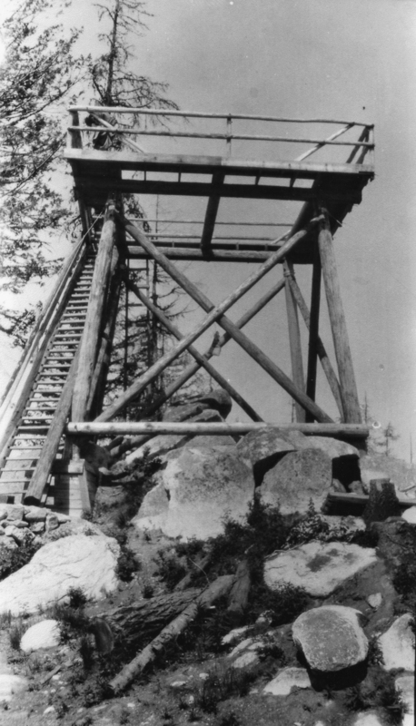 A wooden observation tower with a staircase leading up to a platform, supported by large poles and beams. The structure is surrounded by rocks and sparse vegetation, with trees visible in the background.