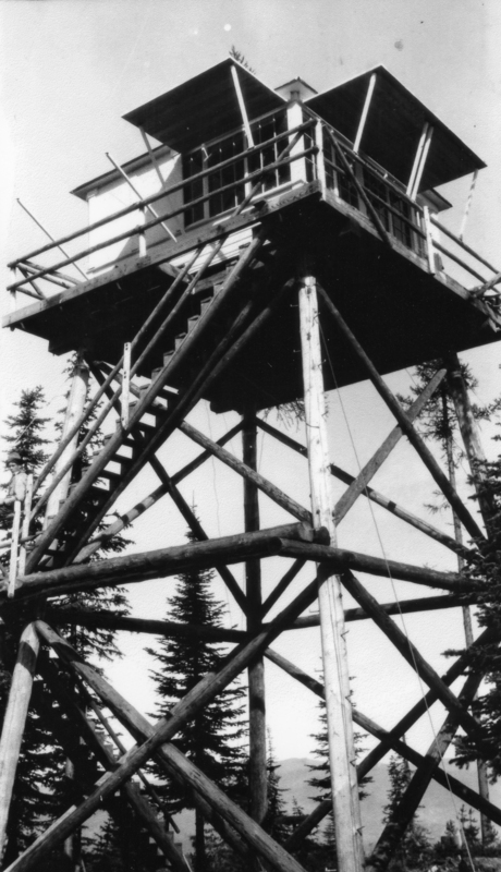 A tall wooden lookout tower with a staircase leading up to an enclosed structure at the top, surrounded by evergreen trees.