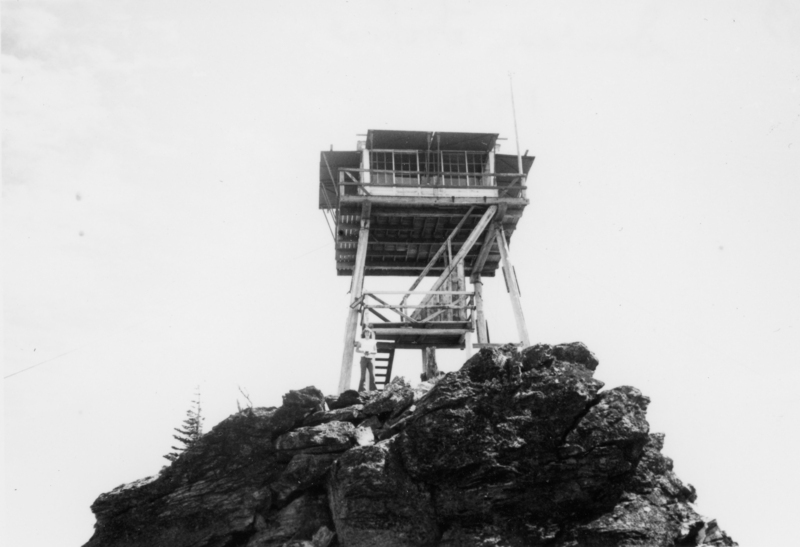 A tall wooden lookout tower stands on a rocky outcrop. A staircase leads up to a platform, where a person is standing, looking at the camera. The structure is supported by several beams.