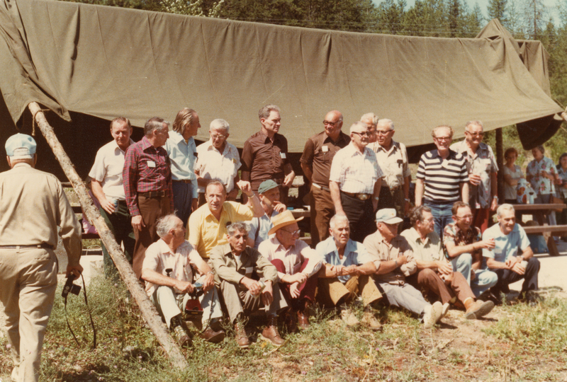 A group of men gathered outdoors, some standing and others sitting on the ground in front of a makeshift tent structure. A man is walking away from the group holding a camera in his hand. Trees are visible in the background.