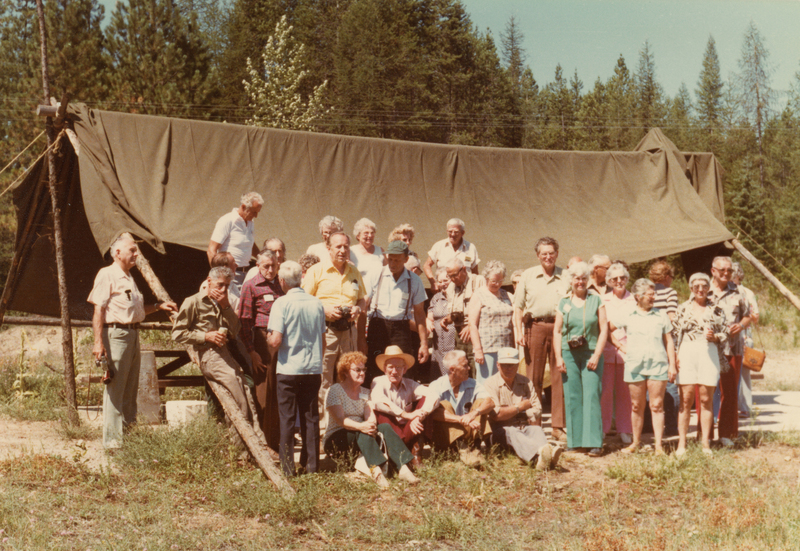 A group of people standing and sitting in front of a large tent in a grassy outdoor area, surrounded by trees. Some individuals hold cameras, and they appear to be casually dressed, possibly attending a gathering or event.