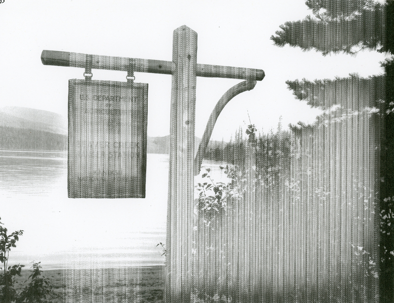A wooden sign hanging from a wooden post near a body of water with trees and shrubs nearby. In the background, there are mountains and a forest.