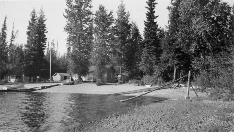 A shoreline with pebbles, a small dock extending into the water, and several buildings including cabins nestled among tall trees.