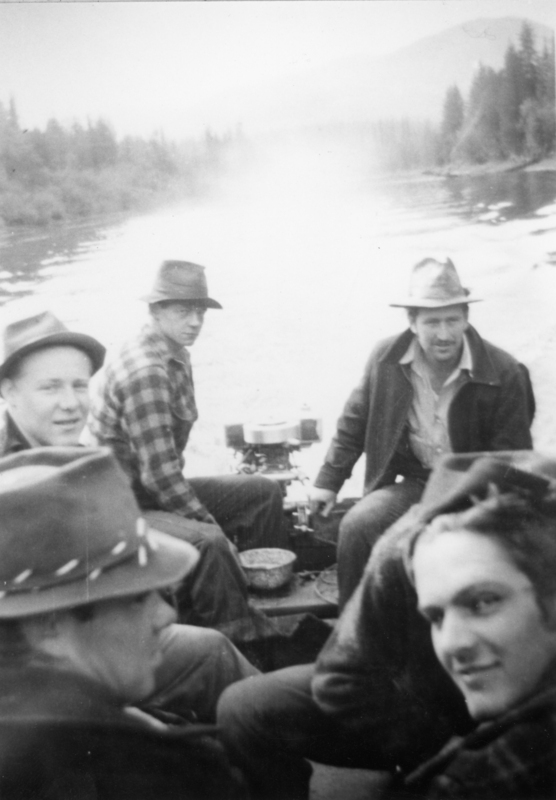A group of men wearing hats are sitting in a boat on a river, surrounded by trees and mountains in the background.