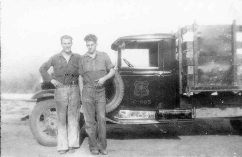Two men standing together in front of a truck with a U.S. logo on the door. The truck has a wooden cargo area, and the men are wearing work clothes. The visible typed text on the side of the truck includes "U.S." inside a shield-like emblem and "C-605" below it.