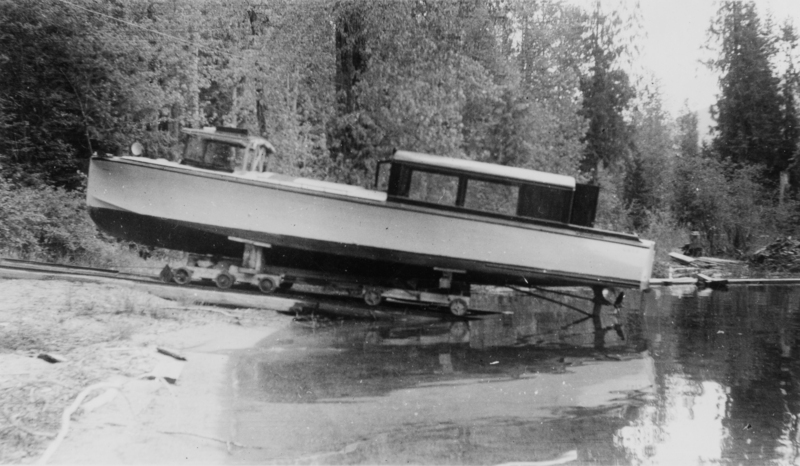 A boat is positioned on a wheeled apparatus, partially submerged in water near a forested area. Trees and foliage surround the scene in the background.