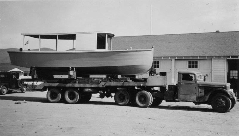 A large boat is loaded on the back of a flatbed truck parked in front of a building with multiple garage doors. A flag is visible at the back of the boat, and a smaller vehicle is parked nearby. The building has a pitched roof, and hills are visible in the background.
