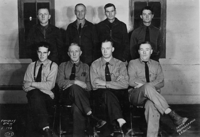 A group of eight men are posed for a portrait. Four men are sitting in the front row with their arms crossed or resting on their laps, while four men stand in the back row. The men are wearing shirts and ties or jackets. Two windows are visible in the background. Text reads: "- Bottom left: "FORESTRY STAFF F. 1 42." - Bottom right: "FORD PHOTOGRAPHIC SERVICE."