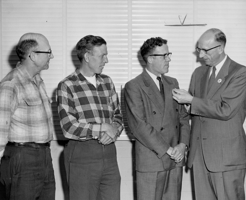 Four men stand in a room with a window behind them. The man on the right is holding something small in his hand and appears to be explaining it to the others. Two of the men are wearing plaid shirts, and two are wearing suits. They are all looking at the object.