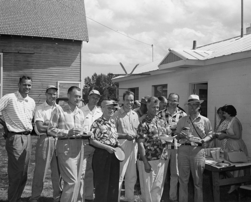A group of men, some holding food and drinks, are standing outside near a table. The men are dressed in casual shirts and pants, and one wears a hat. In the background, two women are near a building, also by a table with cups and boxes. The setting includes a small building with a metal roof and an adjacent wooden structure.