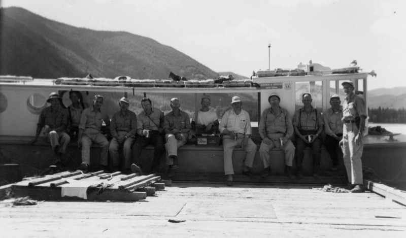 A group of eleven men wearing shirts and hats are sitting and standing on a dock in front of a boat. They are holding mugs and some have boxes. The backdrop features mountains and a body of water. "- Near the top right, on a sign: "DEPARTMENT OF AGRICULTURE" and "FOREST SERVICE." - On a box held by a person: "Rainier." - Near the top right, a logo or emblem of the "FOREST SERVICE.""