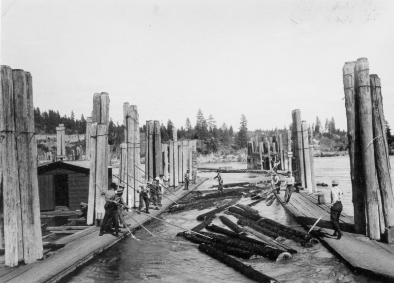 A group of men stand on wooden platforms using long poles to guide logs in a river. Tall wooden posts line the platforms, and there are trees in the background.