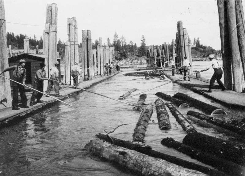 Several men are standing on platforms using long poles to maneuver logs floating in the water. The scene is framed by large wooden pilings, and trees are visible in the background.